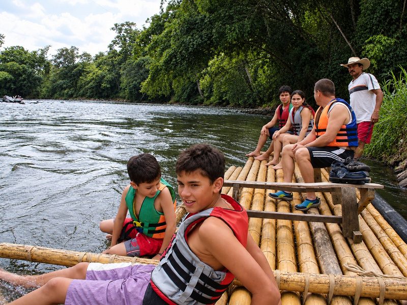 Juanes y Danny en chaleco salvavidas, sentados al borde de una balsa de bambú en el Río La Vieja