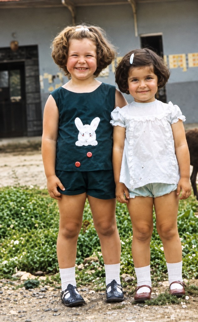 Dos niñas pequeñas de pie, vestidas de verano, sonriendo en un patio de tierra: Alexandra y su hermana en Girardot