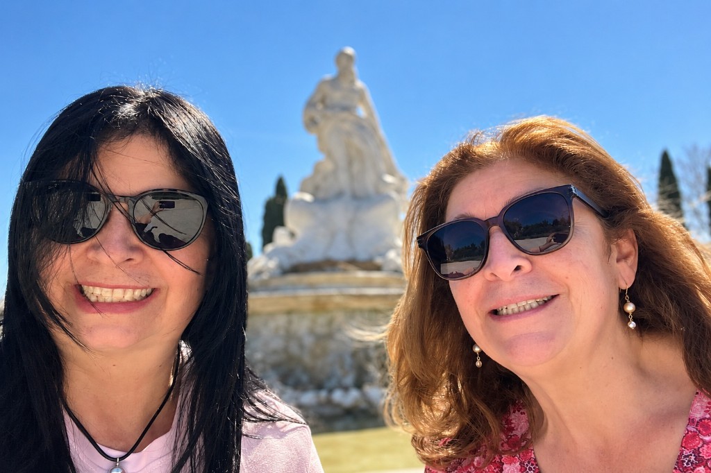 Alexandra y su hermana, con gafas de sol, sonríen juntas frente a una estatua blanca bajo un cielo azul